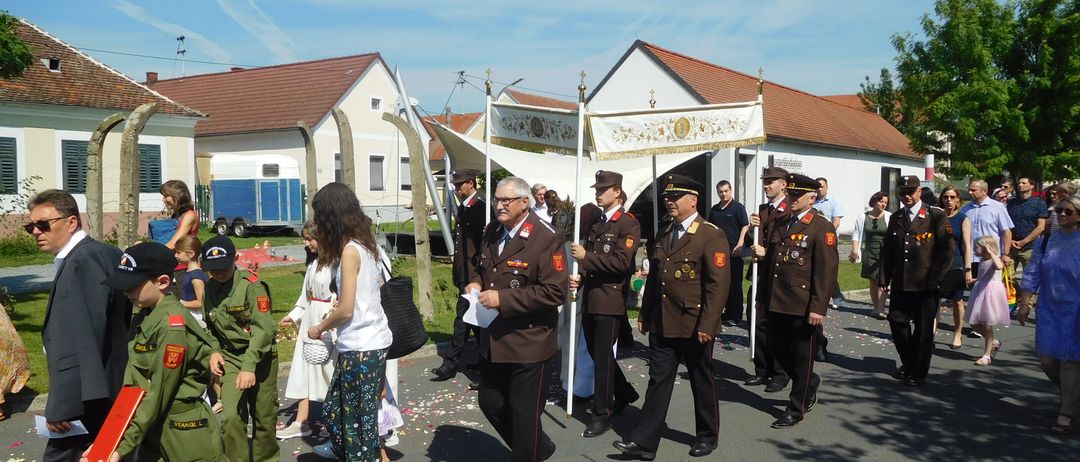 Ein Militäraufmarsch vor einem Haus, Menschen in Uniformen und Kinder in grünen Outfits, einer hält einen Fahnenmast, ein weißes Zelt im Hintergrund und Menschen, die auf einer Straße gehen.