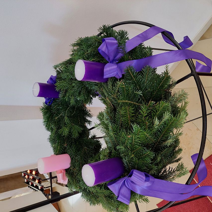 Ein Kranz aus Tannenzweigen mit vier lila Kerzen in einem runden schwarzen Rahmen auf einem gefliesten Boden.
