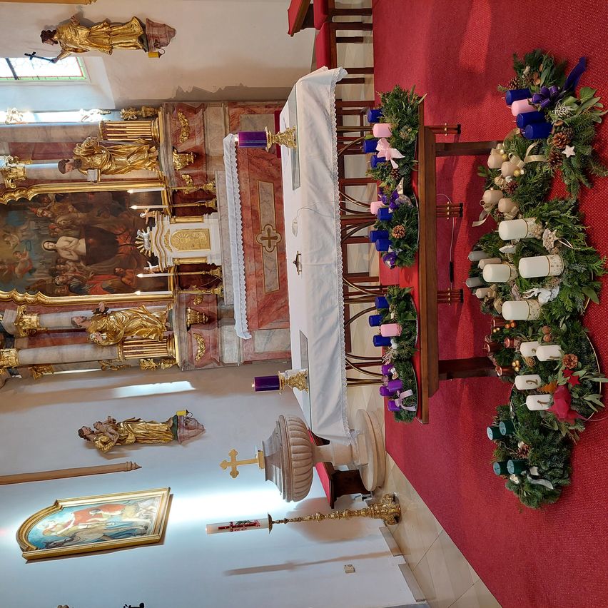 Eine Draufsicht auf eine Kirche mit Altar, Kerzen und goldenen Statuen. Der Altar hat ein weißes Tuch, und Kerzen sind auf beiden Seiten platziert. Eine goldene Statue ist links, und ein roter Teppich liegt auf dem Boden.