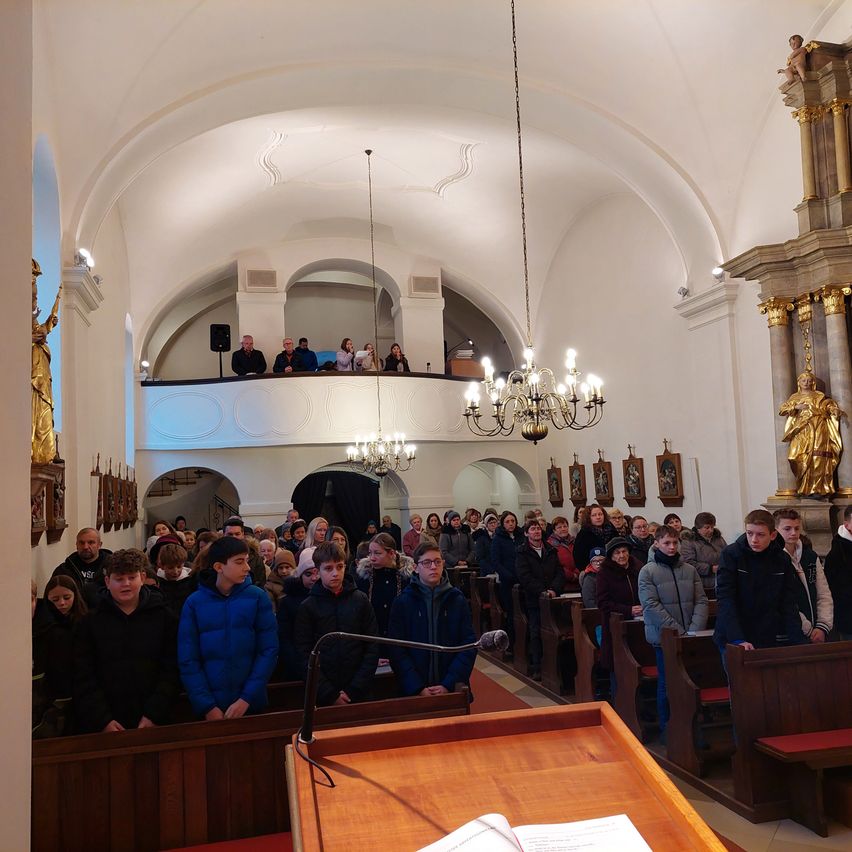 Eine Menschenmenge versammelt sich in einer Kirche für eine Zeremonie, einige stehen und andere sitzen auf hölzernen Kirchenbänken. Der Altar ist mit einer goldenen Statue und Kerzen geschmückt.