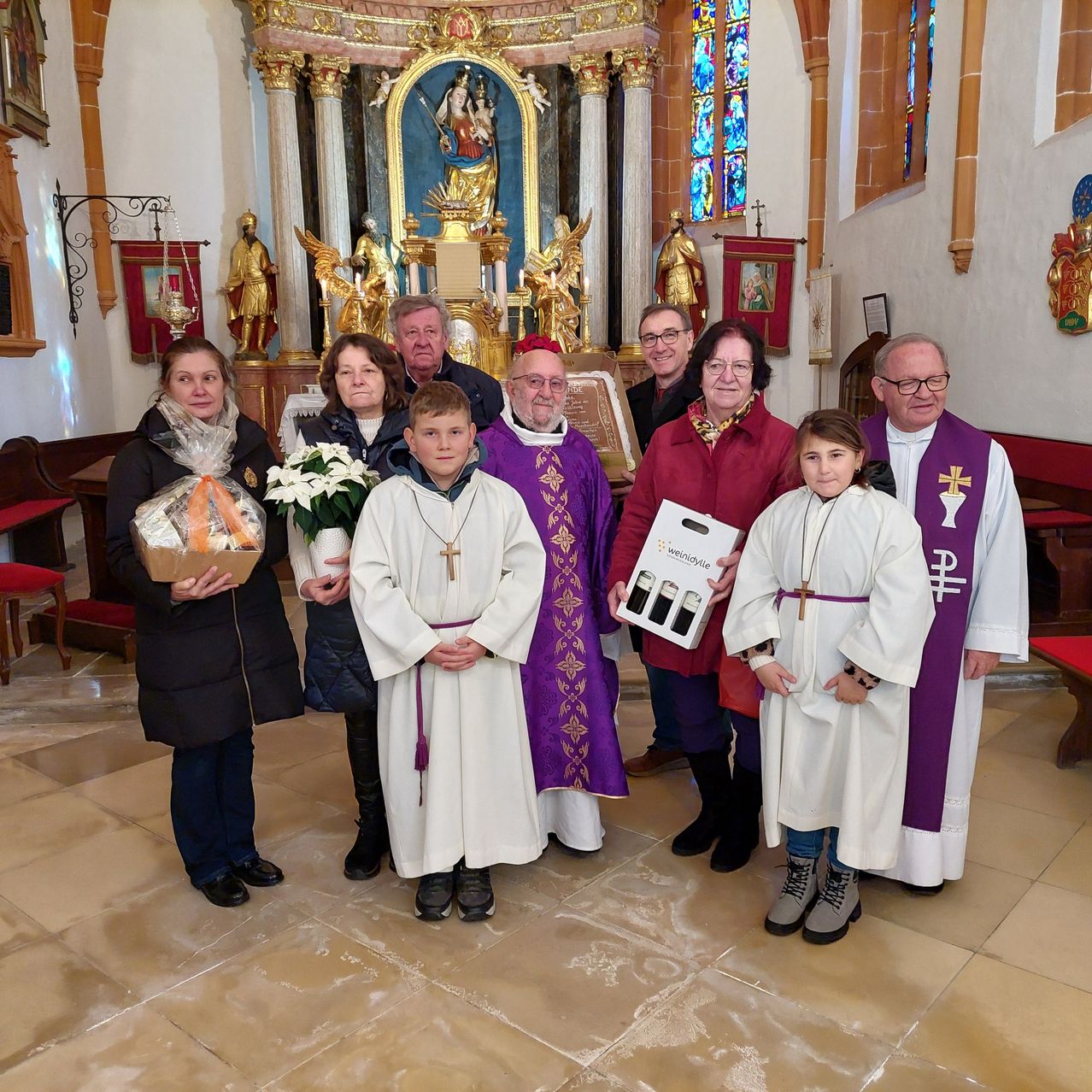 Eine Gruppe von Menschen posiert für ein Foto in einer Kirche. Eine Frau hält einen Korb mit einem Blumenarrangement. Ein Junge und ein Mädchen sind in Weiß gekleidet. Ein Priester hält eine Schachtel.