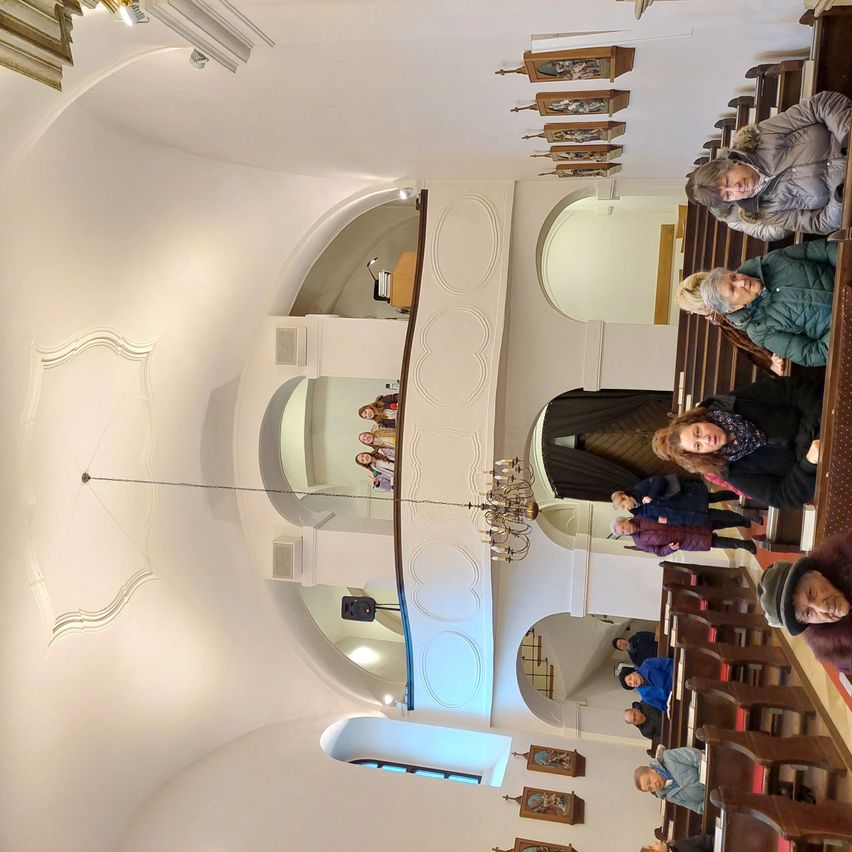 Eine Gruppe von Menschen befindet sich in einer Kirche, einige sitzen auf Bänken, andere stehen, mit einer Kristallleuchte, die von der Decke hängt.