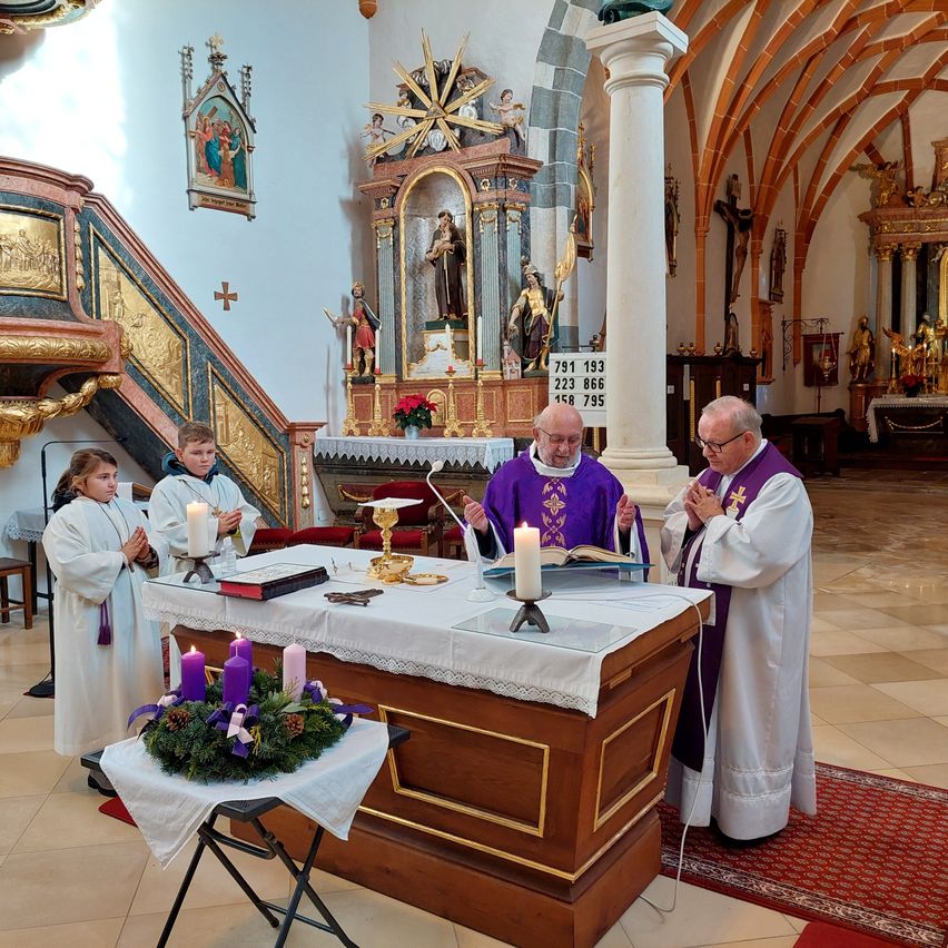 Priester und junge Helfer in weißen Gewändern stehen in einer Kirche, vor einem Altar mit Kerzen und einem offenen Buch. Dahinter sind eine goldene Statue und eine Treppe mit einem Kreuz zu sehen.