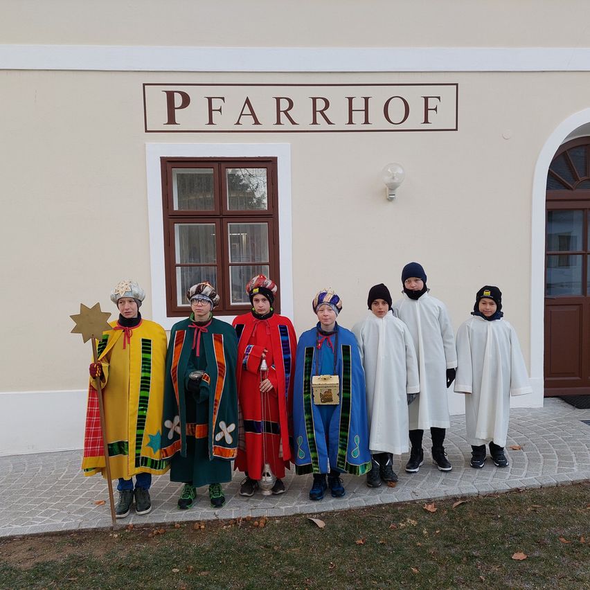 Eine Gruppe von Kindern in traditionellen Kostümen steht vor einem Gebäude mit dem Schild Pfarrhof. Sie posieren für ein Foto.