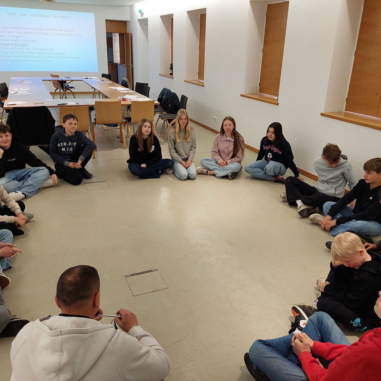 A group of students are sitting in a circle inside a classroom with a projector screen showing a slide. A man is standing in front of them holding a pen.