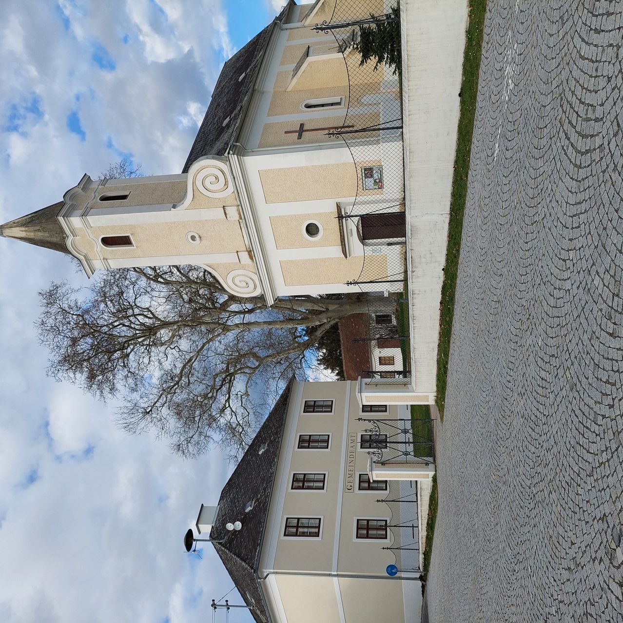 Eine Kirche mit einem Kirchturm und einem Glockenturm. Sie hat ein großes Fenster und ein Kreuz auf dem Dach. Vor der Kirche steht ein Baum.