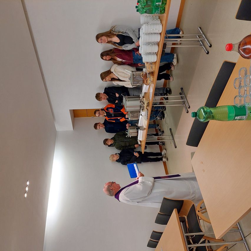 Ein Priester steht vor einer Gruppe junger Menschen am Tisch, mit Flaschen und Gläsern auf dem Tisch.