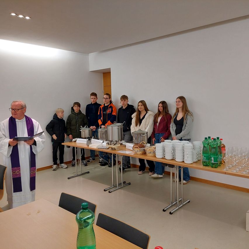 Ein Priester liest aus einem Buch, während eine Gruppe junger Menschen hinter einem Tisch mit Essen und Getränken steht.