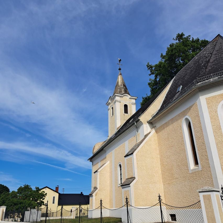 Eine beigefarbene Kirche mit einem Turm und einer Wetterfahne, umgeben von einem Zaun und Bäumen unter einem klaren blauen Himmel.