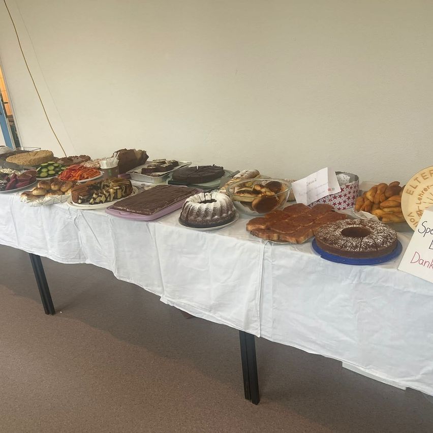 Ein Tisch mit weißer Tischdecke ist mit verschiedenen Desserts wie Kuchen, Donuts und Brot gefüllt. Ein Schild rechts besagt 'Spezieller Dank.' Der Hintergrund ist eine weiße Wand.