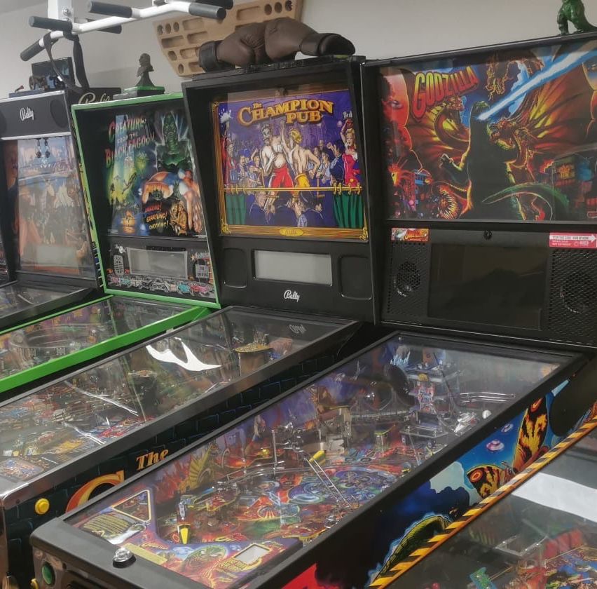 Eine Reihe von alten Arcade-Spielautomaten mit bunten Displays und Glasfronten, darunter Titel wie 'The Champion Pub' und 'Godzilla'.