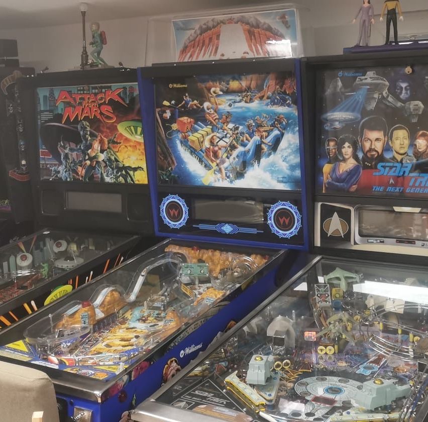 Drei Flipperautomaten sind ausgestellt. Links hat einer ein 'Angriff auf Mars'-Thema, der mittlere zeigt eine dynamische Szene mit Menschen und Fahrzeugen und der rechte hat ein 'Star Trek'-Thema mit Charakteren aus der Serie.