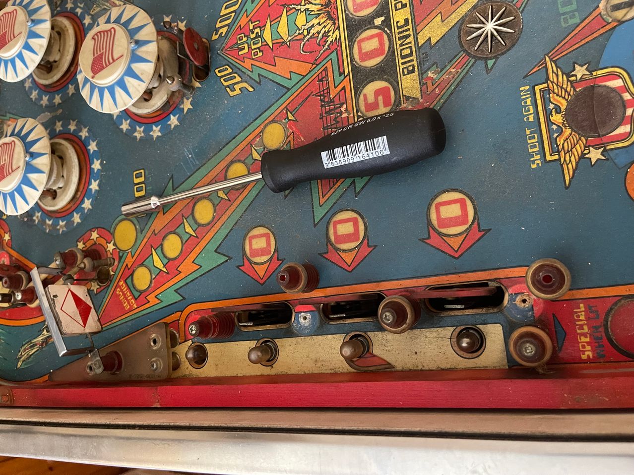 A screwdriver lies on the colorful, worn surface of an arcade pinball machine, surrounded by numerous components and buttons.