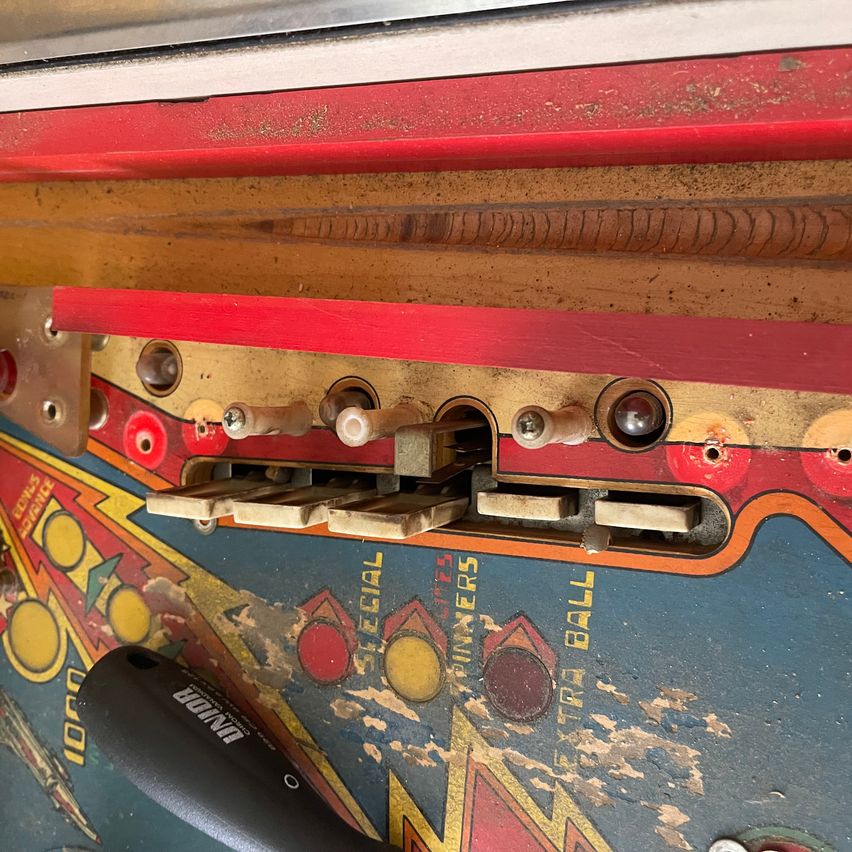 A close-up of an old pinball machine's control panel shows worn-out buttons and switches, with a black handle visible. The machine has a red border and is faded.
