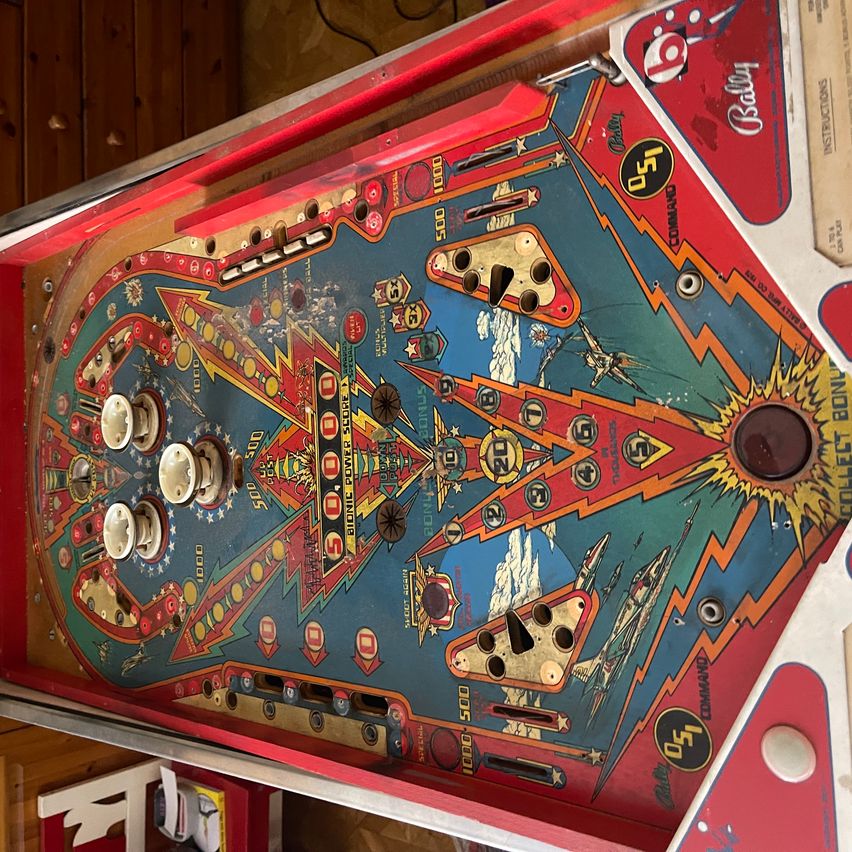 A vintage pinball machine with a blue and red design. It has multiple buttons and holes for gameplay, a black center, and a red border. The machine is placed on a wooden surface.