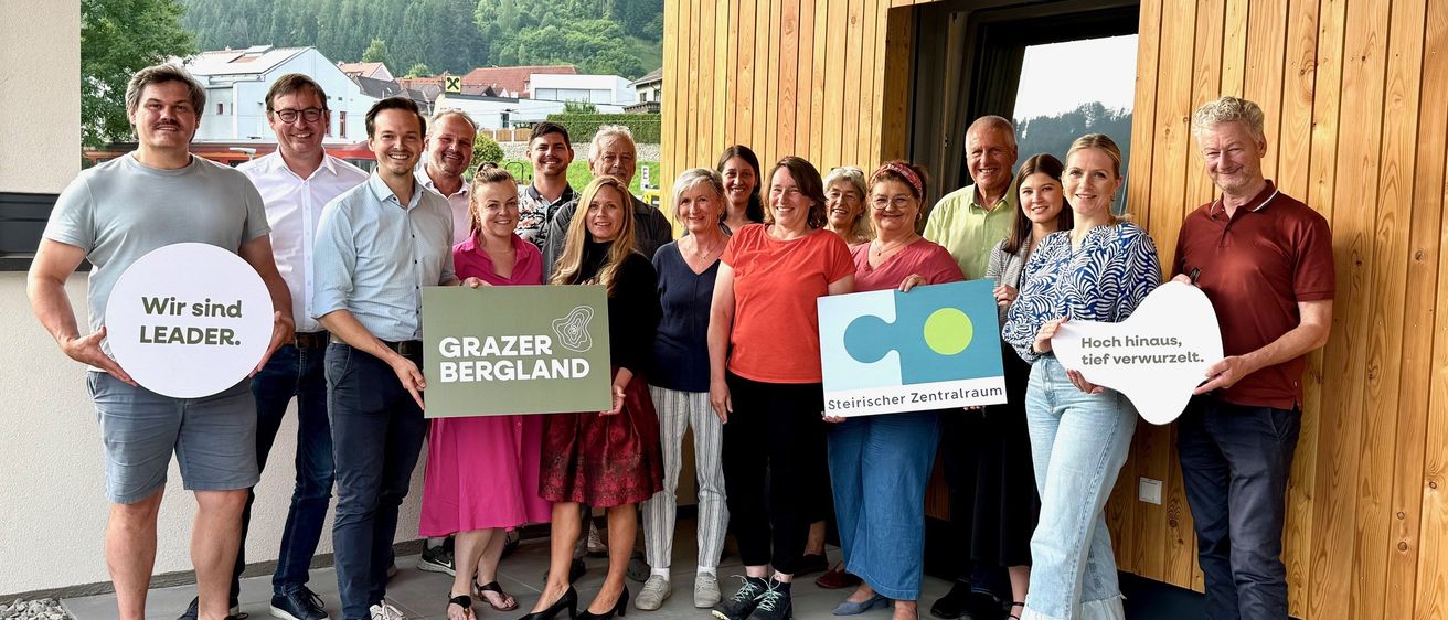 Eine Gruppe von Menschen steht vor einem Holzgebäude, hält Schilder und posiert für ein Foto. Im Hintergrund sind ein Berg und Gebäude zu sehen.