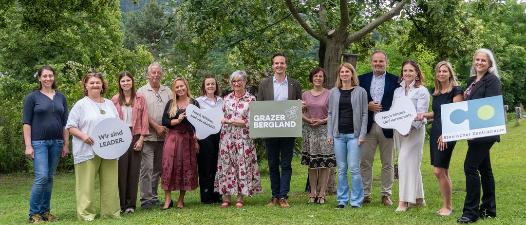 Eine Gruppe von Menschen steht in einem Park und hält Schilder mit den Aufschriften 'Wir sind LEADER' und 'Grazer Bergland'. Sie posieren für ein Foto.