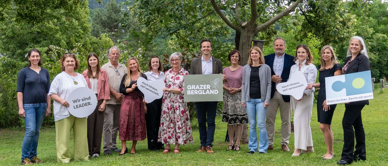 Eine Gruppe von Menschen steht in einem Park und hält Schilder mit den Aufschriften 'Wir sind LEADER' und 'Grazer Bergland'. Sie posieren für ein Foto.