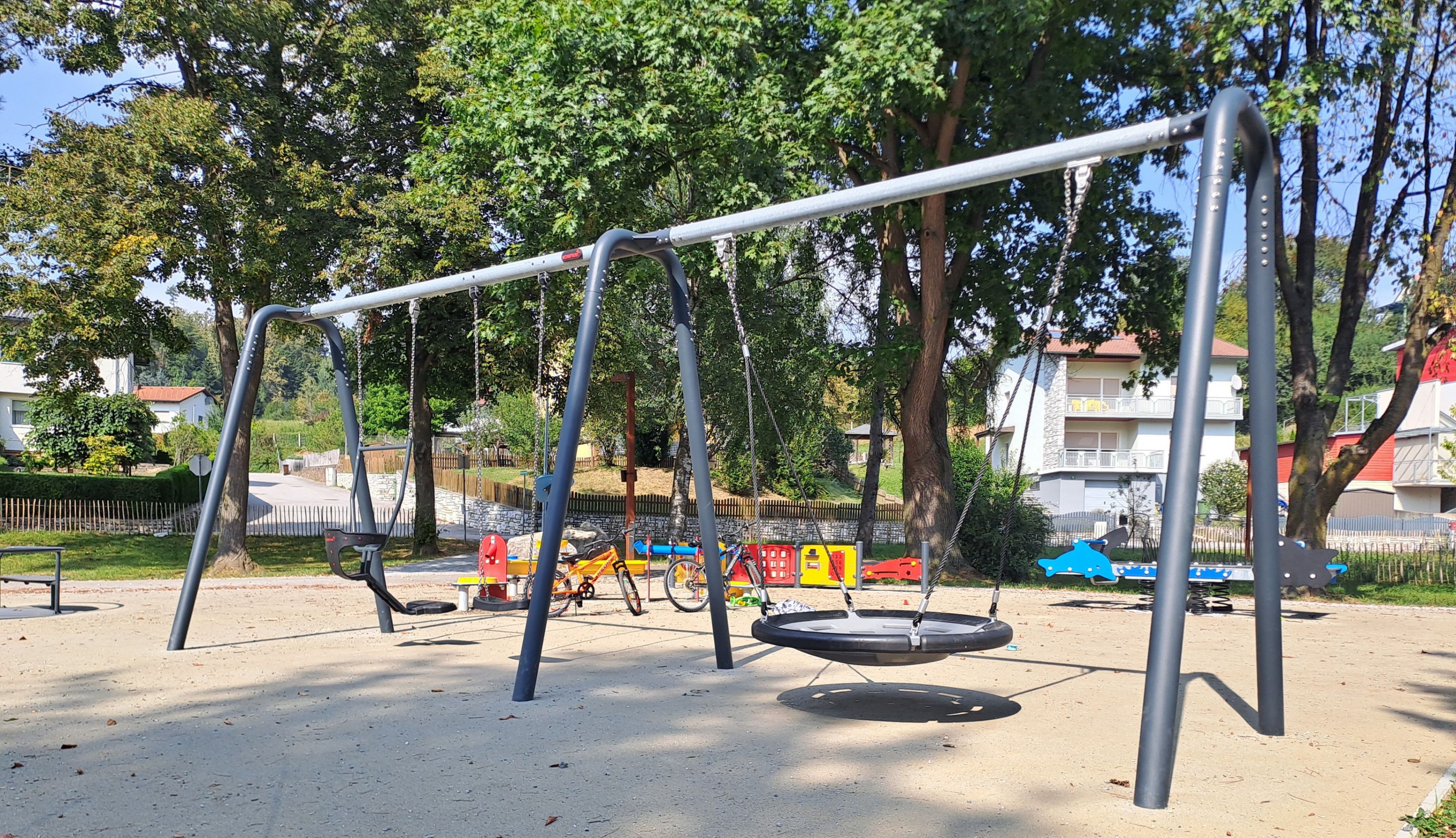 Ein Spielplatz mit zwei Schaukeln, einer Reifenschaukel und einem Fahrrad, das in der Nähe eines Baumes geparkt ist. Es gibt auch bunte Spielzeuge und ein Gebäude im Hintergrund.