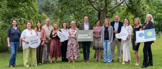 Eine Gruppe von Menschen steht in einem Park mit einem Schild, das Grazer Bergland sagt. Sie alle lächeln und scheinen für ein Foto zu posieren.