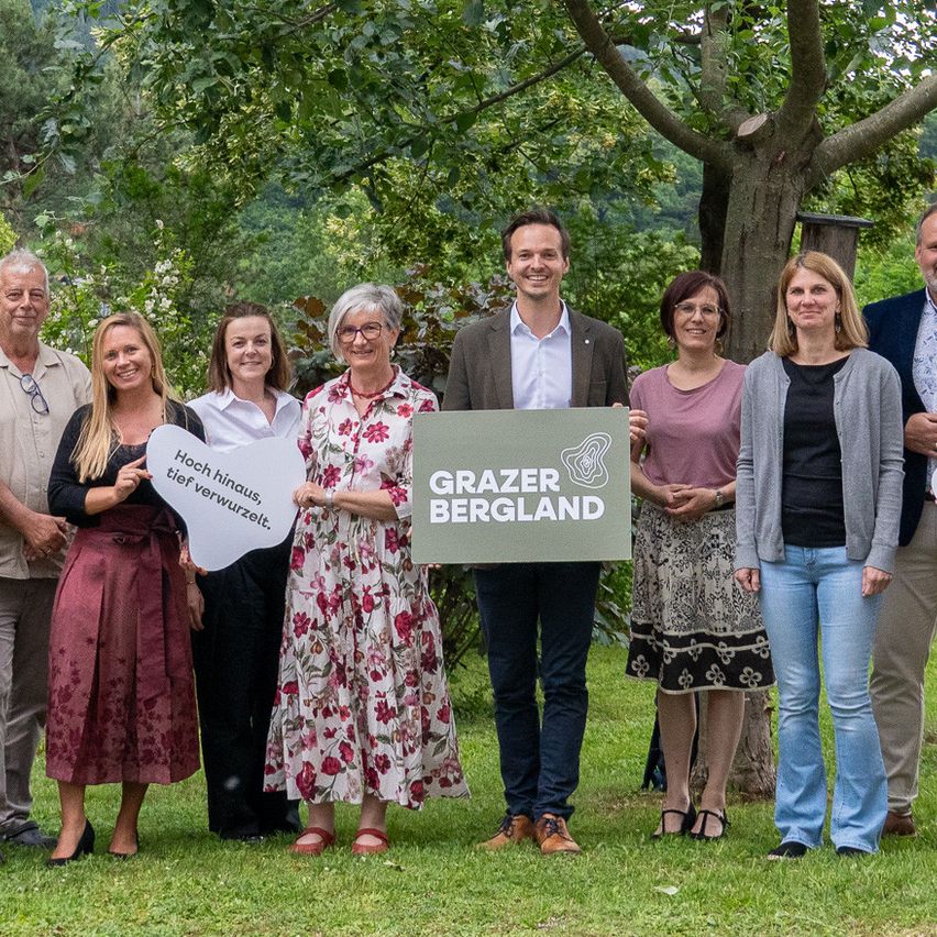 Eine Gruppe von Menschen steht in einem Park mit einem Schild, das Grazer Bergland sagt. Sie alle lächeln und scheinen für ein Foto zu posieren.