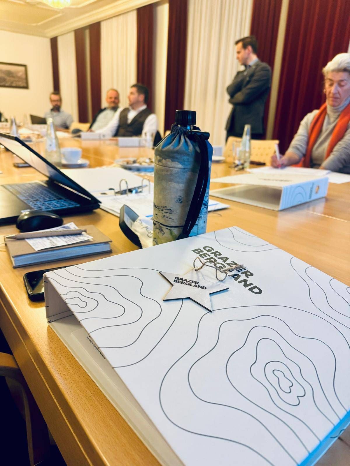 Ein Besprechungstisch mit Laptop, Buch, Wasserflasche und Notizbuch. Ein weißer Sternabzeichen ist an einer Mappe mit dem Titel 'Grazerbergland' befestigt. Drei Personen sitzen am Tisch.