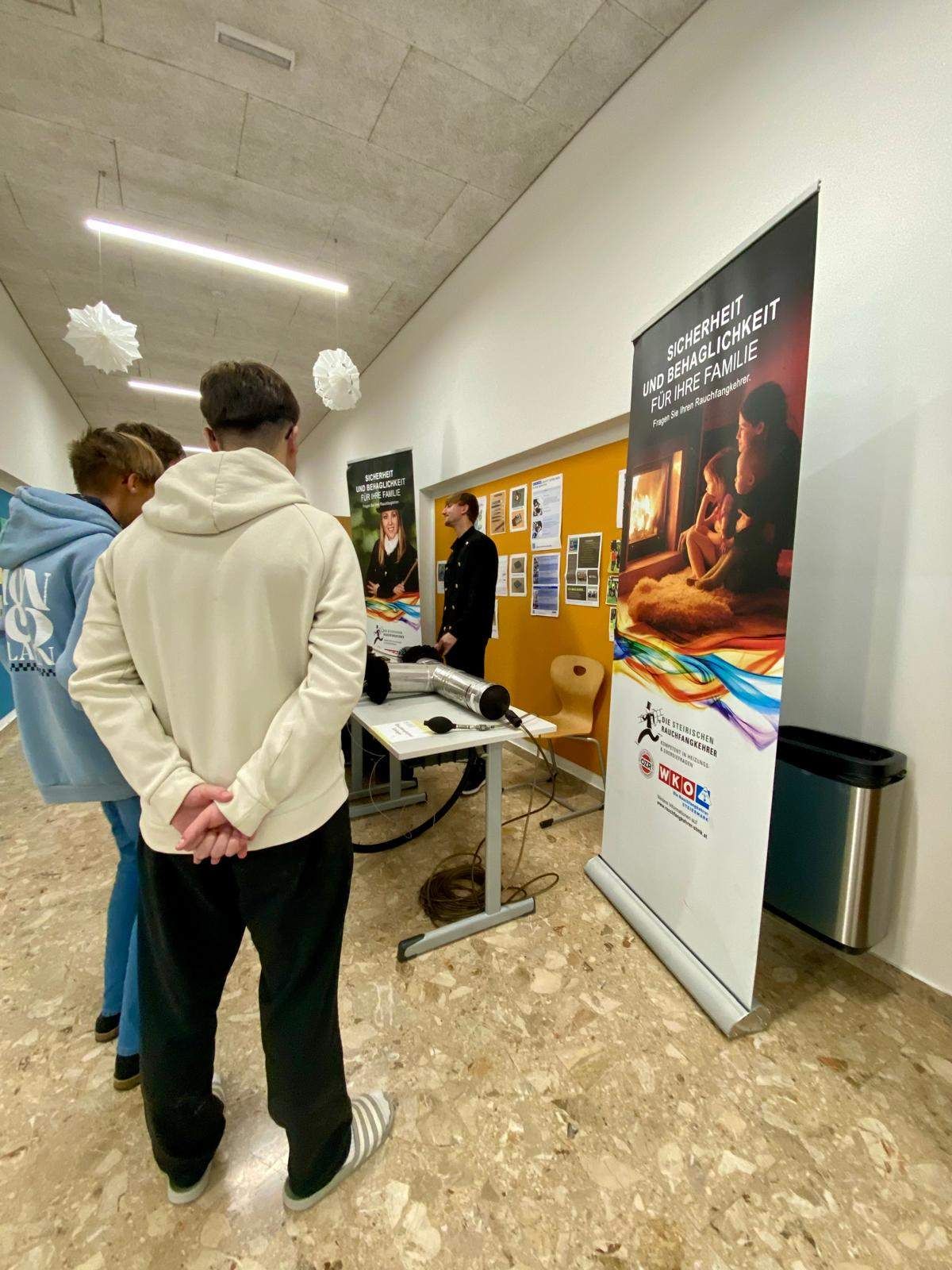 Vier Personen stehen und betrachten einen Stand mit einem Poster über Sicherheit und Glück für die Familie. Der Stand befindet sich in einem Flur mit gelber Wand und weißer Decke.