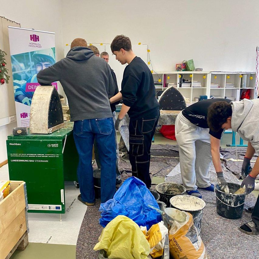 Eine Gruppe junger Männer arbeitet in einem Raum an einem Projekt. Einer gießt Zement in einen Eimer, während andere in der Nähe stehen. In der Nähe befindet sich ein Banner mit einem Bild eines Mannes.