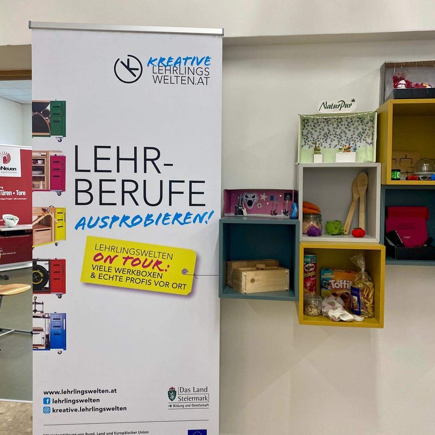 Ein weißer Banner, der für eine Tour durch Lehrberufe wirbt. Helle gelbe und blaue Akzente unterstreichen die Botschaft. Im Hintergrund sind verschiedene farbenfrohe Regale mit Gegenständen und Kisten zu sehen.