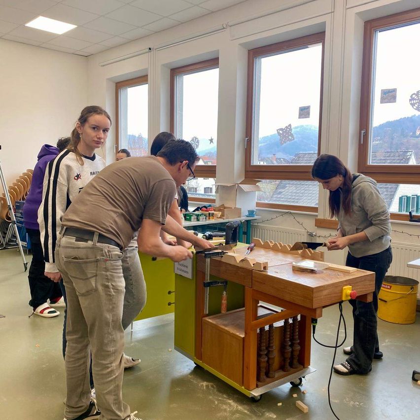 Eine Gruppe von Menschen arbeitet an einem Holzarbeitstisch in einer Werkstatt. Ein Mann benutzt eine Säge, während eine Frau in der Nähe ihre Arbeit konzentriert beobachtet. Eine Frau und ein Mann stehen daneben und beobachten. Der Raum hat große Fenster mit Blick auf die Berge.