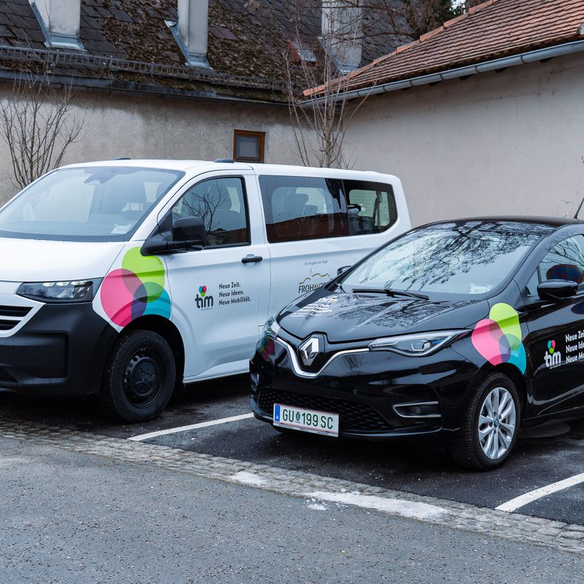 Two cars are parked on the side of the road, one in front of the other. The white van has a colorful design on the side, and the black car has a license plate GU 199 SC.