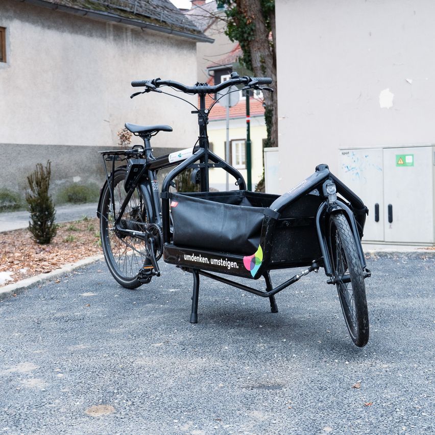 A black cargo bicycle is parked in a lot. It has a basket in front and a bag attached to the rear. The bicycle is parked near a building with peeling paint.