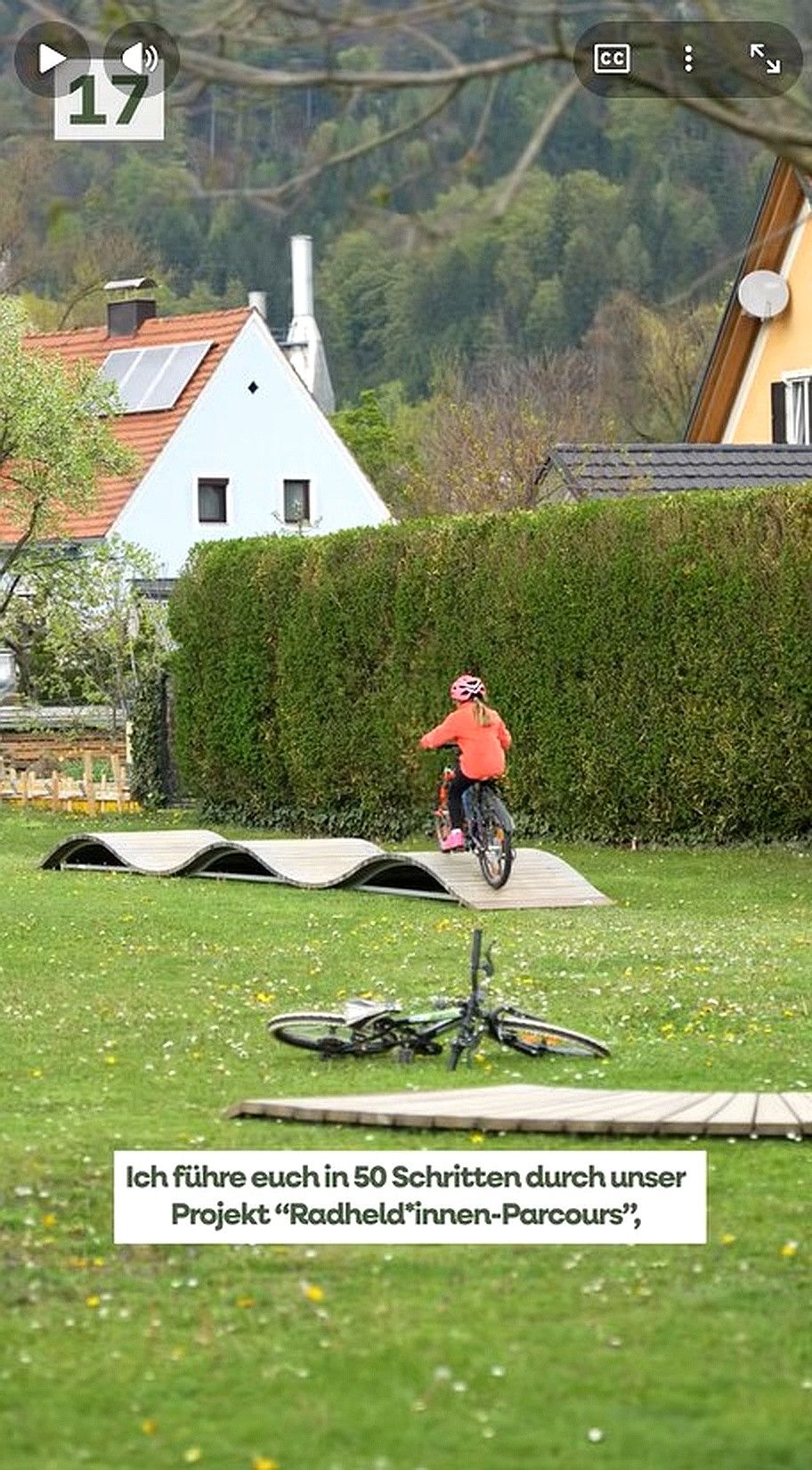 Ein Kind fährt mit dem Fahrrad über eine Holzrampe im Park, mit Häusern und Solarpanelen im Hintergrund.