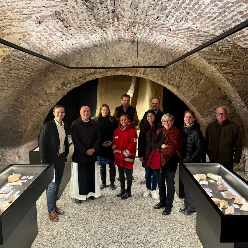A group of people stands in a vaulted room with brick walls, smiling for a photo. Some wear winter clothing, and others wear glasses. Display cases with rocks are in the foreground.
