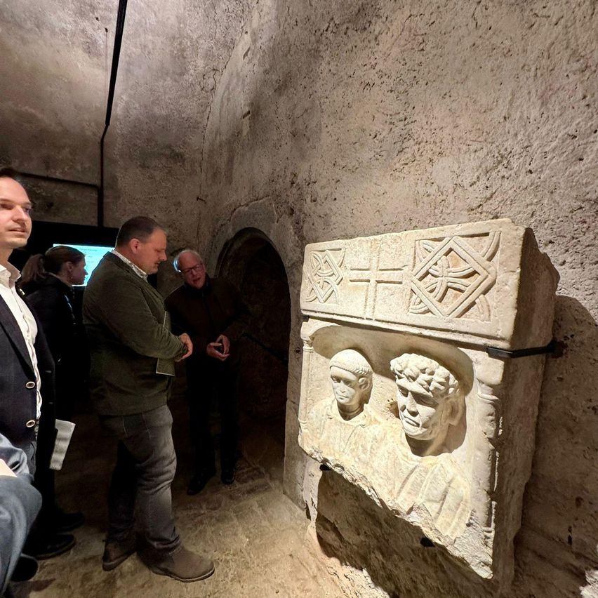 A group of people are standing in a dimly lit room with stone walls, looking at a stone sculpture depicting two men.