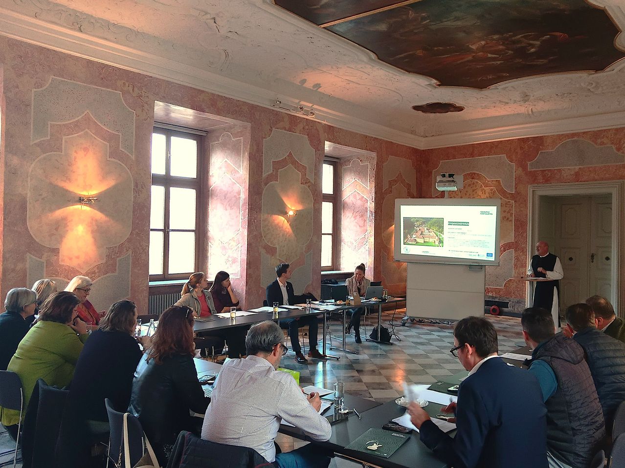 A group of people sit at a table in a conference room, looking at a projector screen. A man stands at a podium, and others are seated and standing around the room.