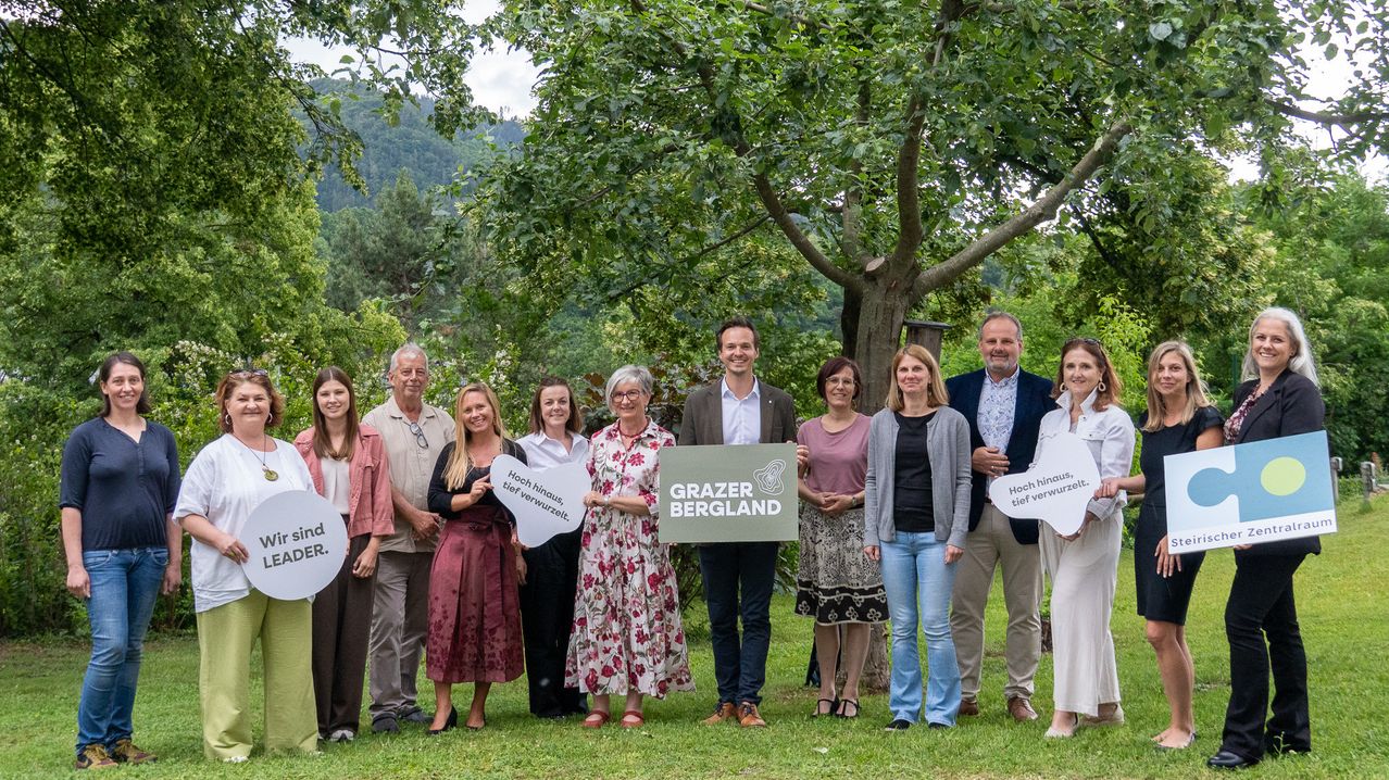 Eine Gruppe von Menschen steht in einem Park mit einem Schild, das 'Grazer Bergland' besagt. Sie halten Schilder mit der Phrase 'Hoch hinaus, tief verwurzelt'. Der Hintergrund zeigt Bäume und einen Berg.