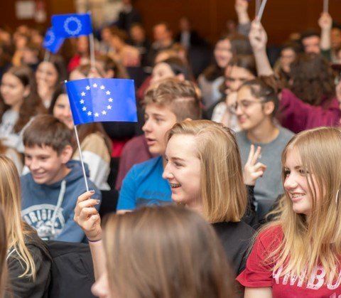 Eine Gruppe junger Menschen in einem Hörsaal, einige halten Flaggen der Europäischen Union hoch, wirken enthusiastisch und engagiert.
