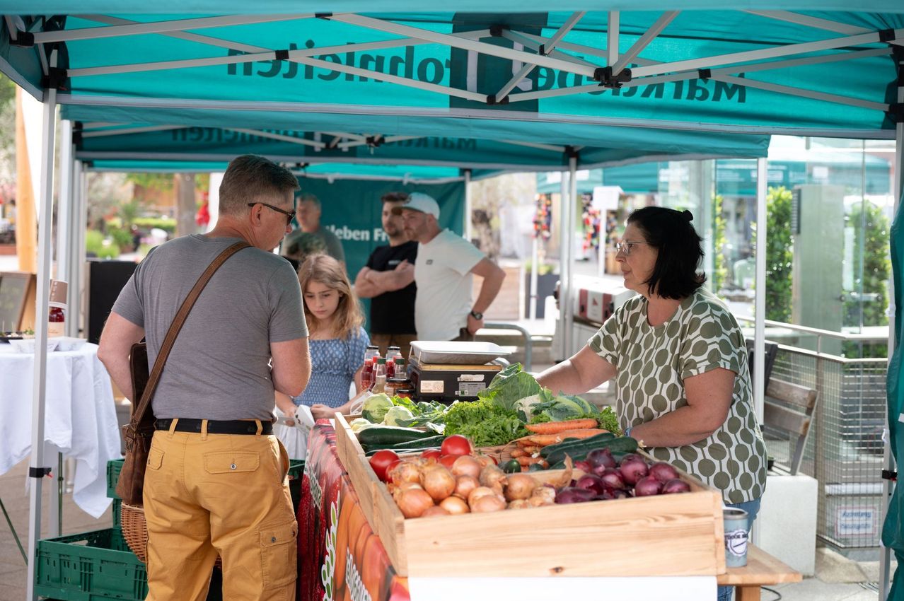 Eine Frau verkauft Gemüse unter einem blauen Zelt auf einem Markt. Ein Mann und ein Mädchen kaufen Gemüse.