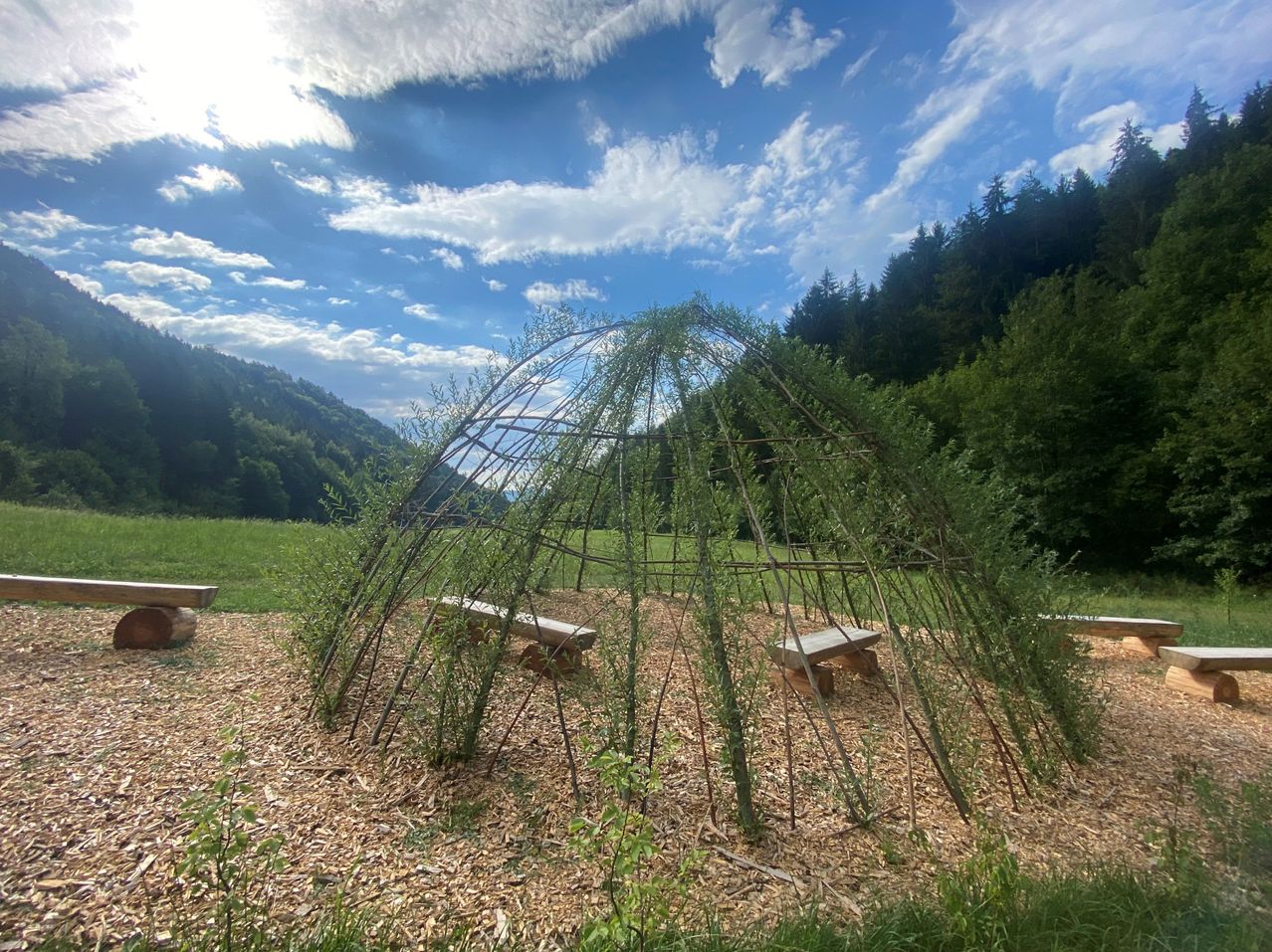 Eine rustikale Holzstruktur, die einem kleinen Hütten- oder Unterstand aus ineinander verflochtenen Ästen ähnelt, in einem Grasfeld mit einem Hintergrund aus Bergen und Bäumen unter einem blauen Himmel mit Wolken.