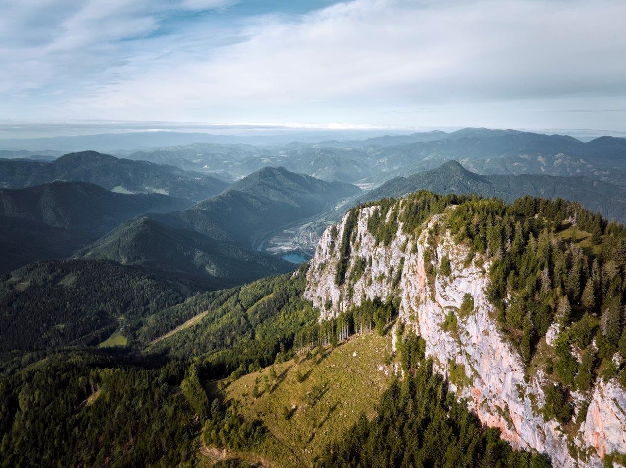 Aerial view of a mountainous landscape with dense forests, a rocky cliff, and a river snaking through the valley. The sky is partly cloudy.