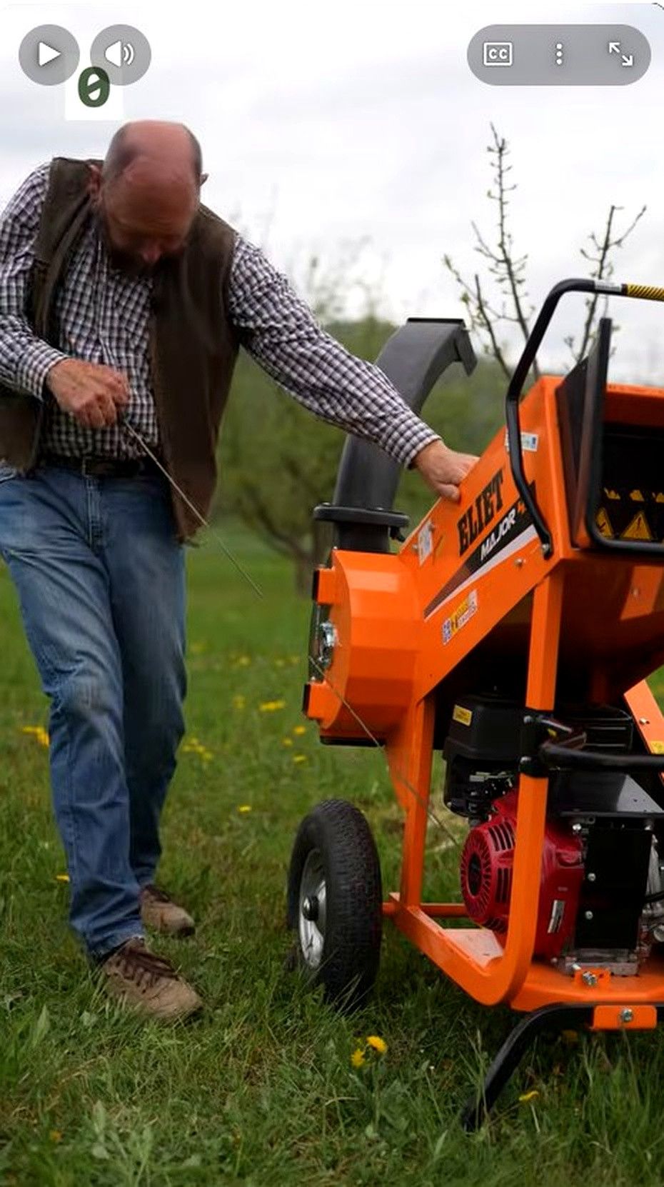 A man in a plaid shirt and vest operates an orange wood chipper in a grassy field.