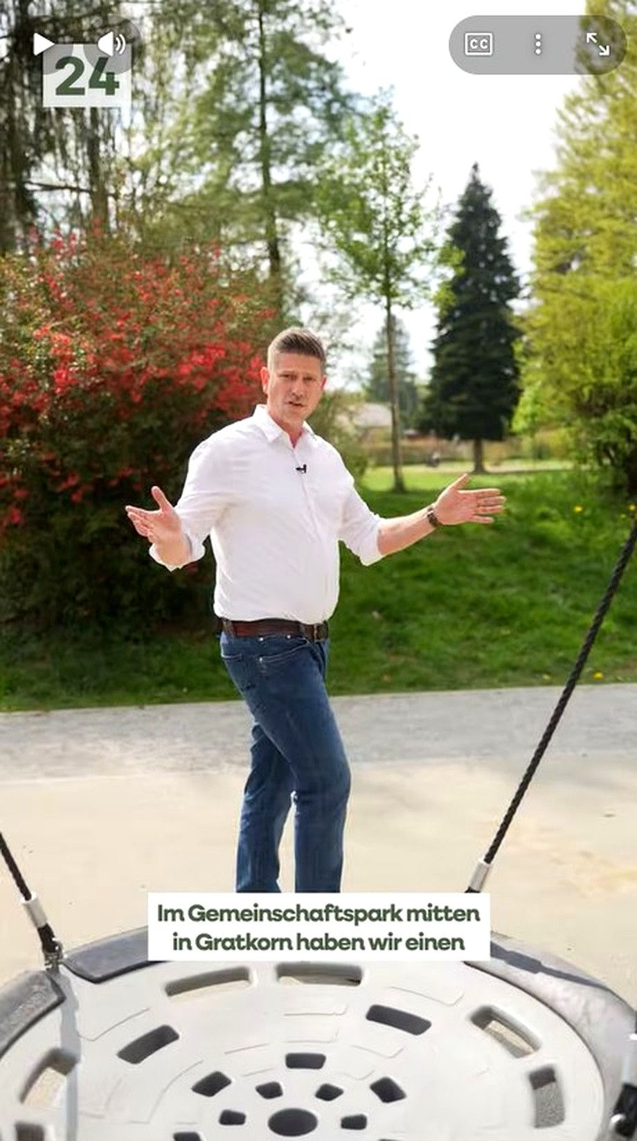A man stands in a park, gesturing with his hands and wearing a white shirt and blue jeans.