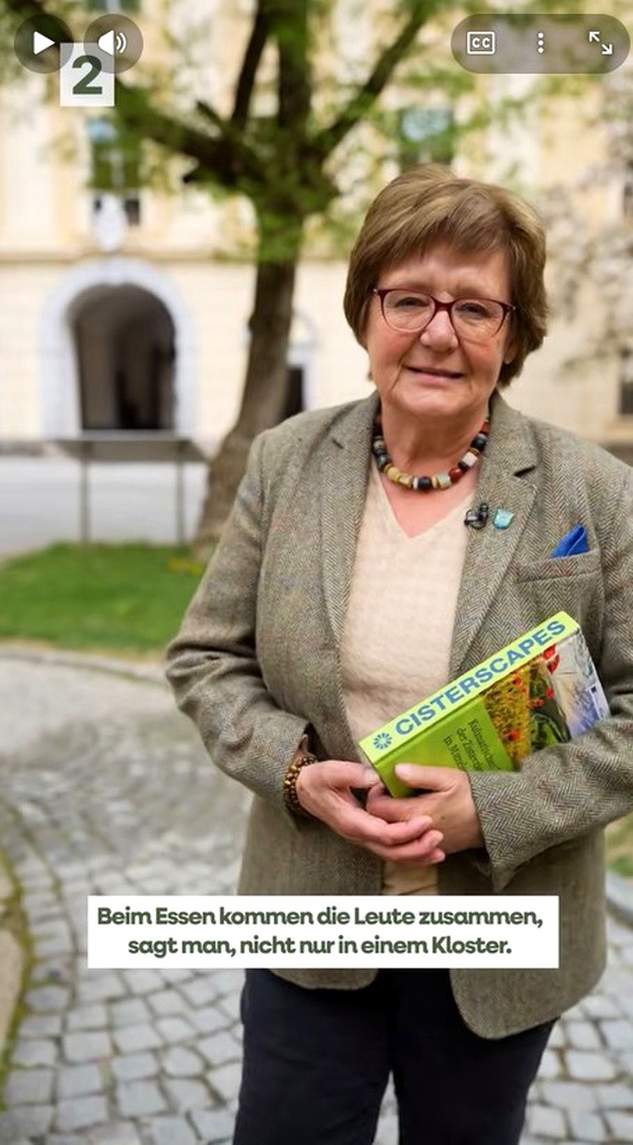 Eine Frau mit Brille steht draußen und hält ein Buch mit dem Titel 'Cisterscapes'. Sie trägt einen grauen Blazer und eine Halskette. Dahinter steht ein verschwommenes Gebäude und ein Baum.
