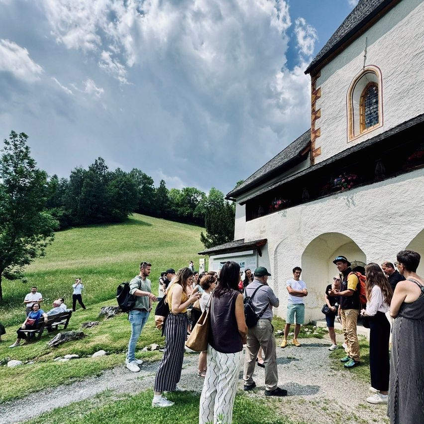 Eine Gruppe von Menschen steht vor einer Kapelle, einige mit Rucksäcken. In der Nähe sitzen andere auf einer Bank unter einem Baum. Im Hintergrund ein Hügel und ein bewölkter Himmel.