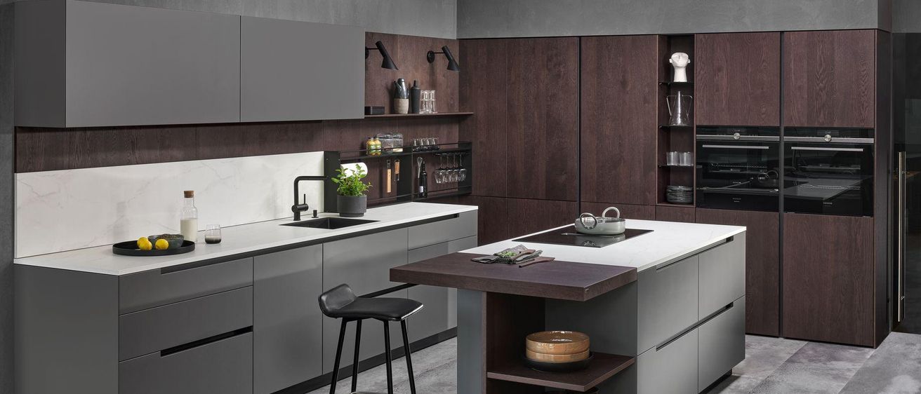 Eine moderne Küche mit Holzschränken und Arbeitsplatten. Ein schwarzer Barstuhl steht in der Ecke, nahe einem Waschbecken mit Wasserhahn. Auf der Arbeitsplatte befinden sich ein Teekanne und andere Gegenstände.