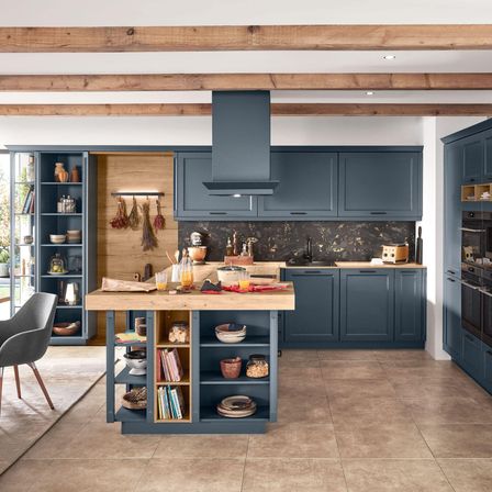 Eine moderne Küche mit Holzinsel, blauen Schränken und einer Marmorarbeitsplatte. Kräuter hängen über dem Herd, und die Küche wird von Deckenleuchten beleuchtet.