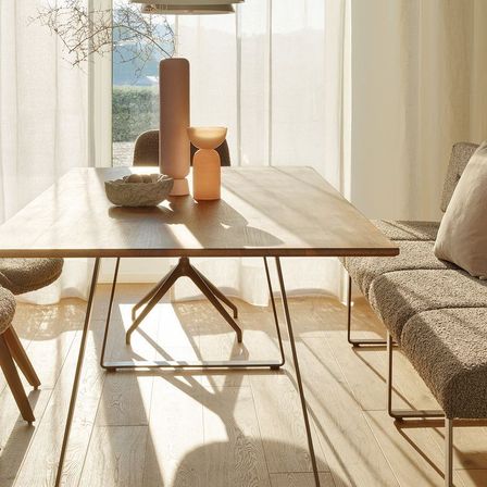 Ein modernes Esszimmer mit einem Holztisch und Stühlen. Ein beiger Sofa mit Kissen steht in der Nähe. Der Raum hat einen minimalistischen Stil mit klaren Linien und neutralen Farben.