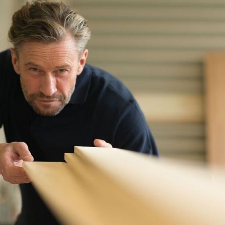 Ein Mann in einem dunkelblauen Shirt untersucht Holzbretter und konzentriert sich auf eines mit seinen Händen.