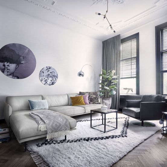Ein modernes Wohnzimmer mit einer weißen Couch, Kissen und einem Couchtisch. Ein großer Teppich, eine Pflanze und dekorative Lampen sorgen für Wärme. Zwei runde Kunstwerke und ein Stuhl vervollständigen den Raum.